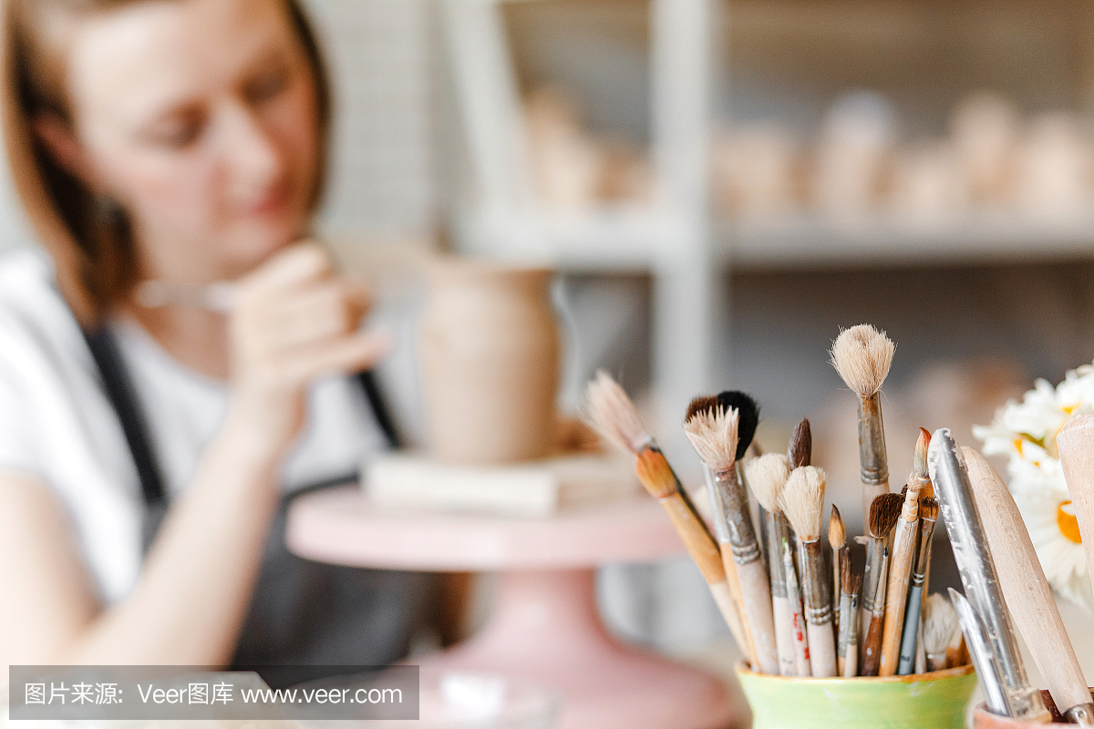 在现代化的车间里,一个女艺术家穿着围裙微笑着在画笔和其他工具之间绘制陶土